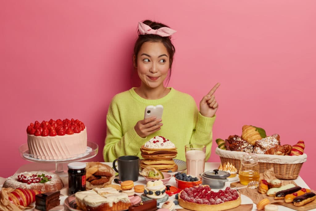 cheerful glad brunette asian girl surrounded by biscuits cookies cakes enjoys sweet food during festive time enjoys holidays treats points blank space uses mobile phone